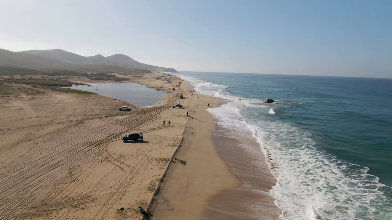 Aerial view of cars parked on a sandy beach beside ocean surf and an inland water area.
