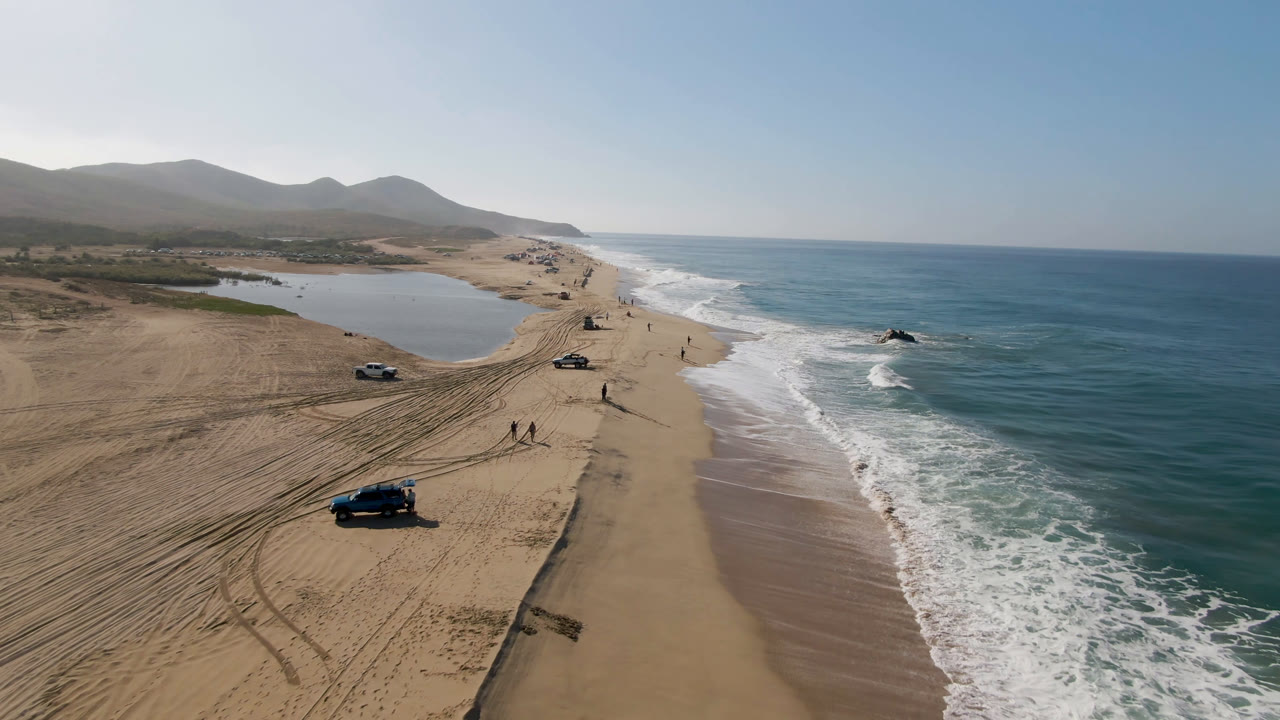 Aerial frame from Migrino, Baja California Sur, showing a dark vehicle nearer the lower beach, other cars along the sand, white surf advancing at right, inland water at left, and small distant people visible.