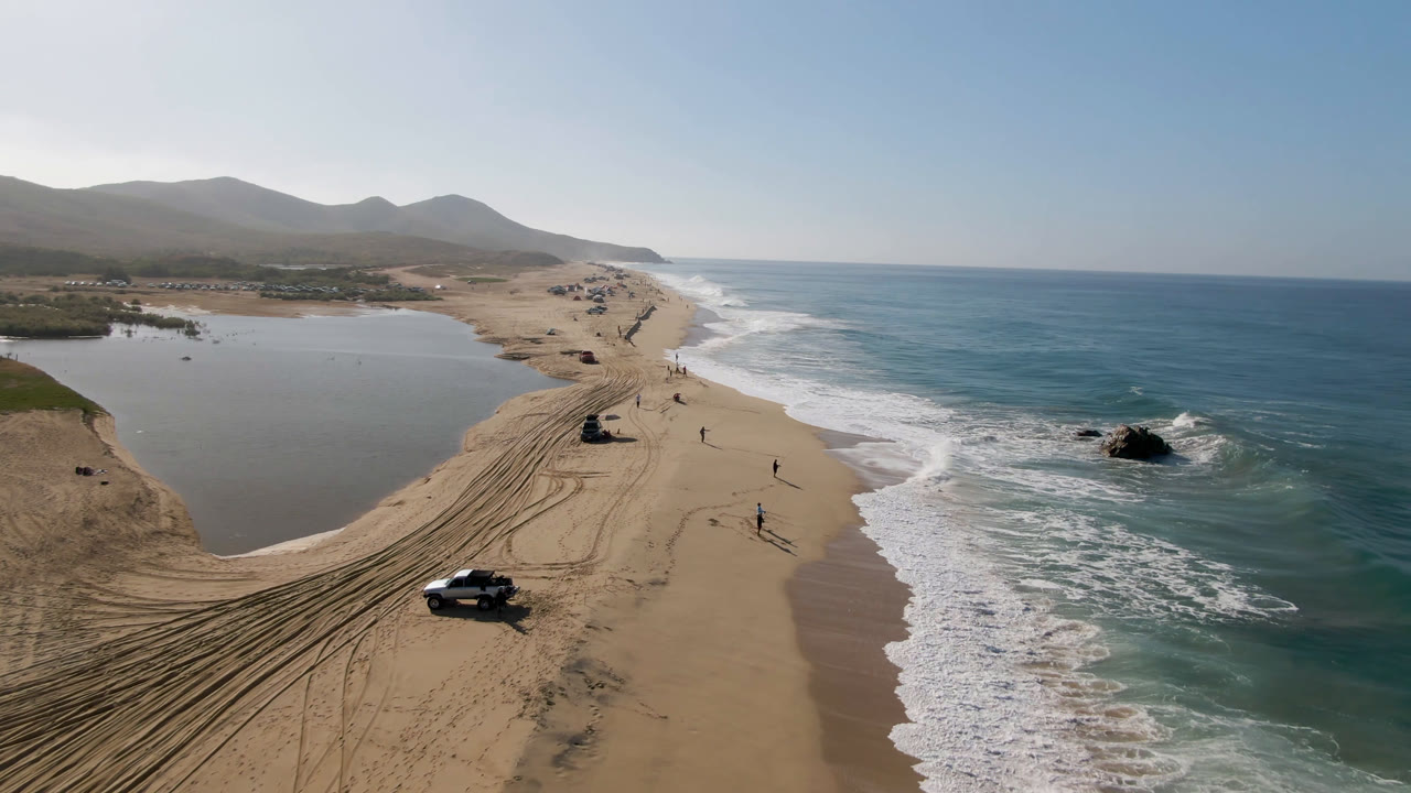 Drone view of sandy beach between lagoon and ocean with vehicles and surf.