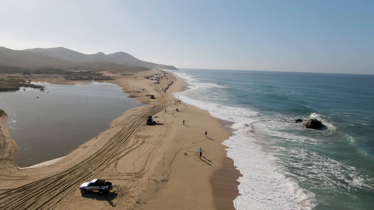 Drone frame over Migrino beach with a white vehicle near the lower sand tracks, lagoon water on the left, ocean surf on the right, small walkers along the shoreline, and mountains beyond the beach.