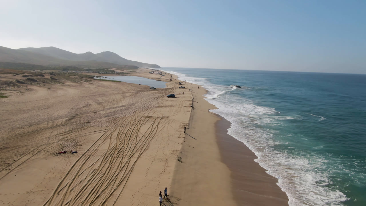 Aerial view over Migrino beach in Baja California Sur, Mexico, showing tire-marked sand, parked cars near the shoreline, white surf, a small inland water area, and distant mountains at the start of the clip.