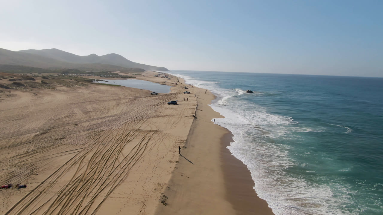 Drone view of Migrino shoreline with a broad tan beach, several vehicles spaced along the sand, surf breaking beside the beach, and hazy mountains in the background.