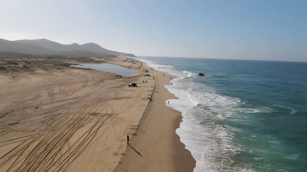Aerial coastline with parked cars on sand, breaking waves, and a rocky point offshore.