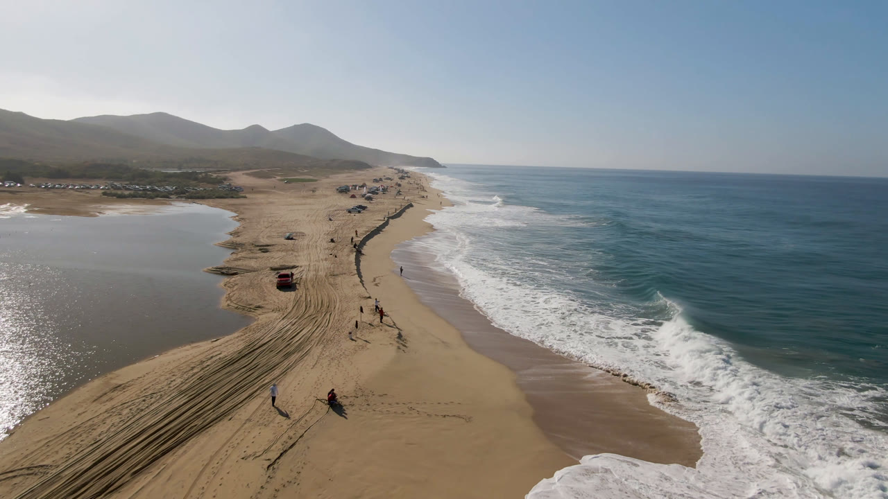 Aerial view of a sandy beach with surf, lagoon water, vehicle tracks, and vehicles in a line.