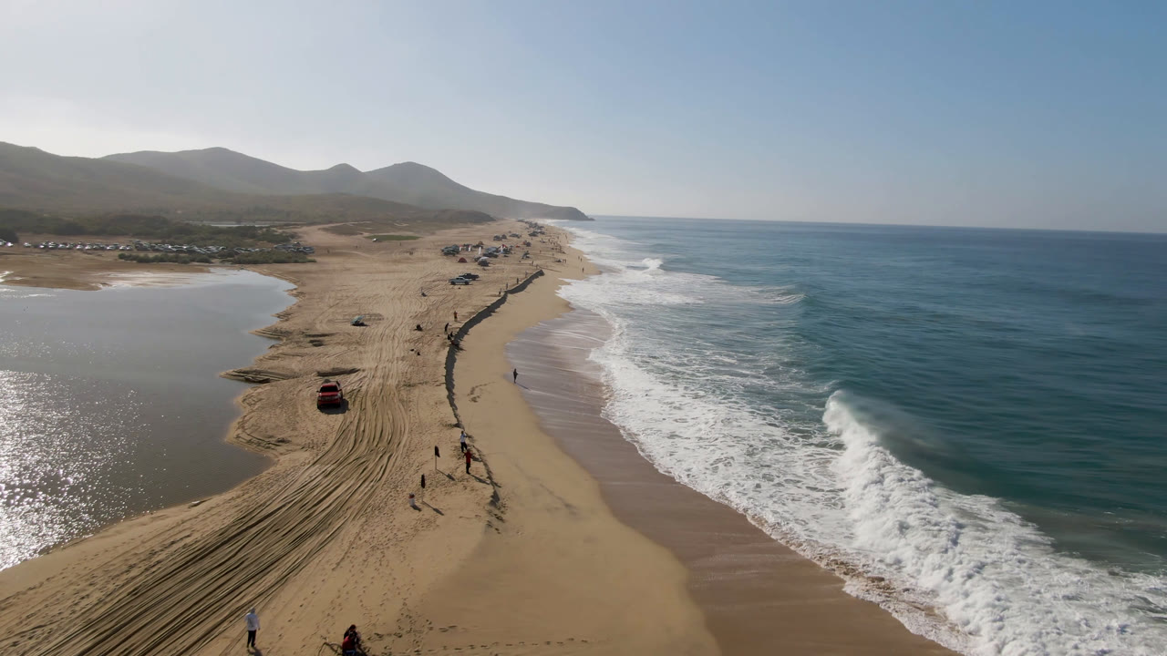 Aerial frame over Migrino beach showing the red vehicle nearer the lower sand, a long track corridor through the beach, breaking surf, and hazy mountains beyond.