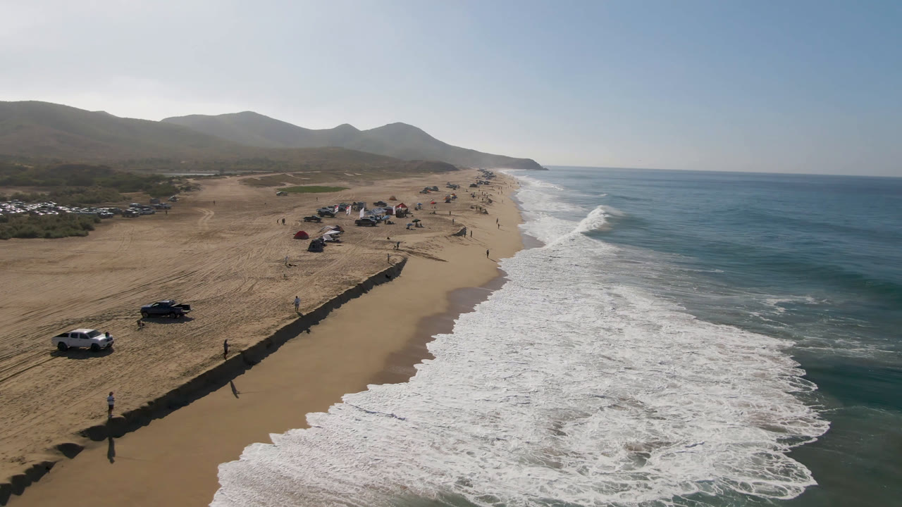 Aerial view of sandy beach, surf, parked vehicles, and distant hills.