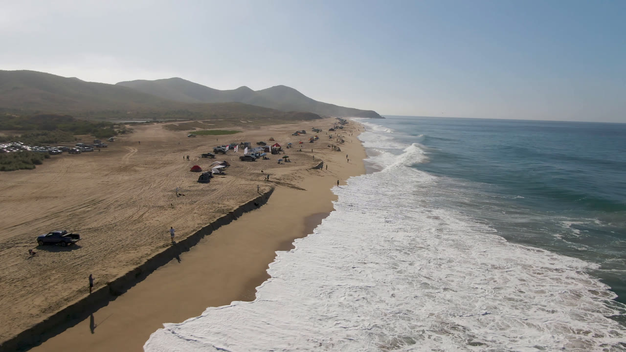 Migrino beach seen from above with the shoreline angled through the frame, foamy waves on the right, vehicles and shade setups on the sand, and small people walking near the water.