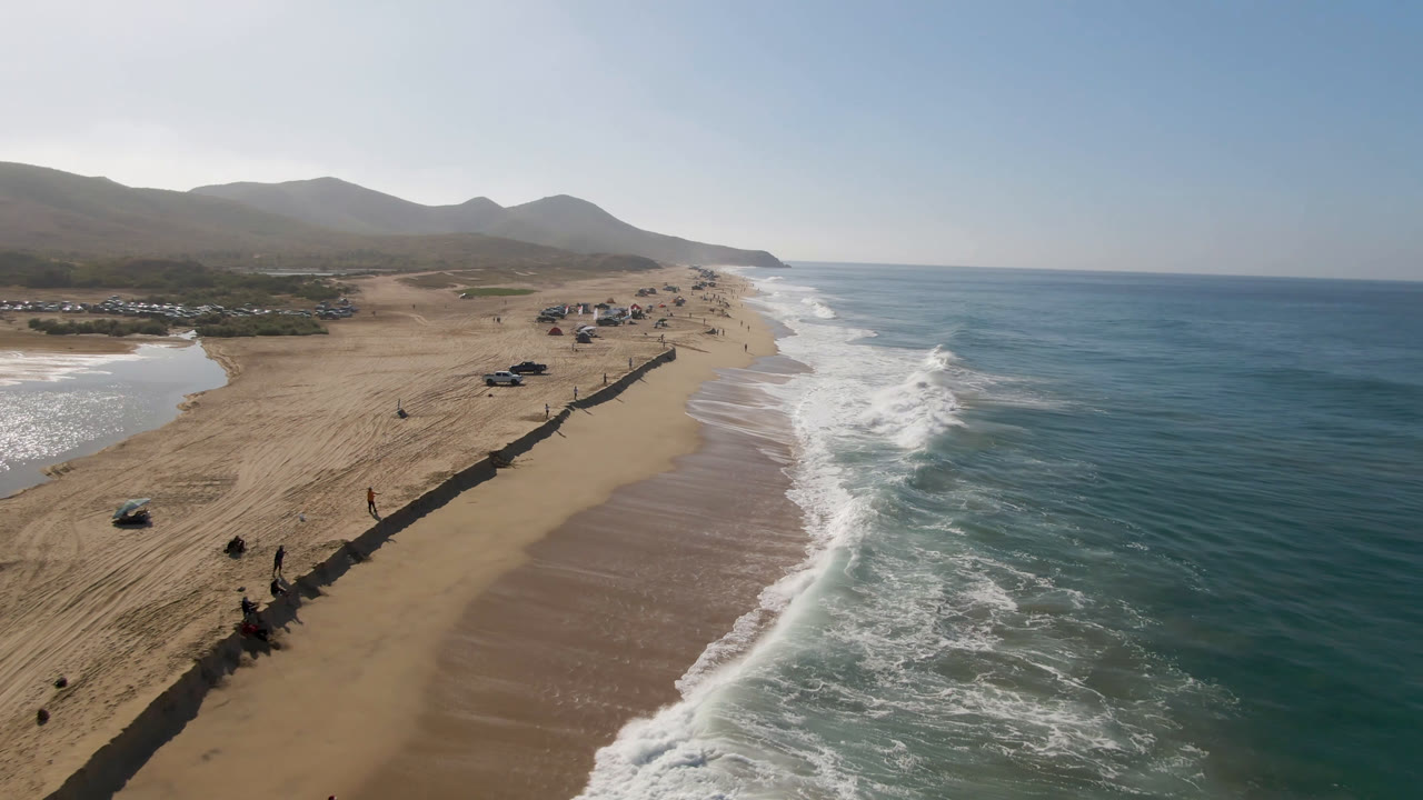 Aerial beach view with surf, parked cars, sandy flats, and mountains.