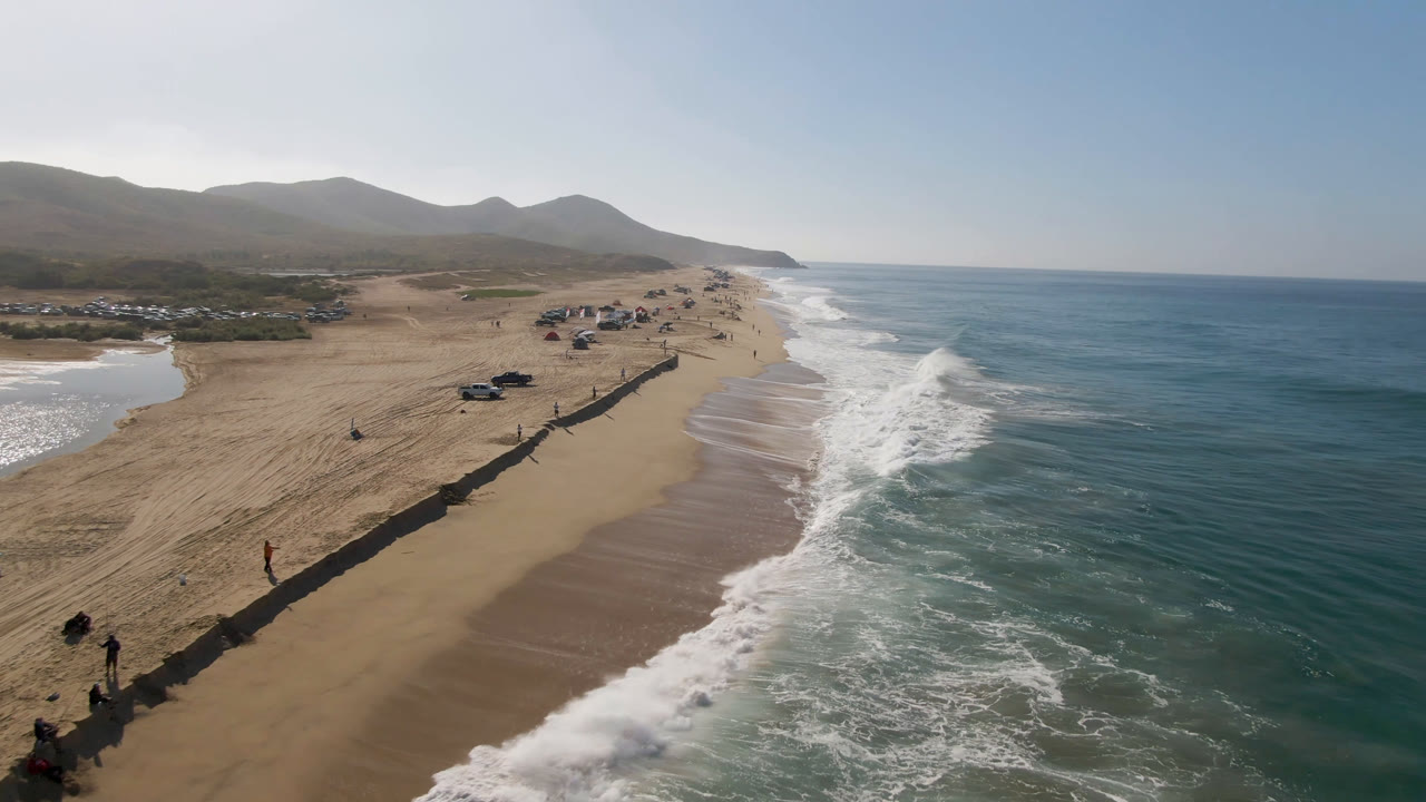 Drone view over Migrino beach with parked vehicles near a long dark barrier, foamy waves reaching the shoreline, mountains in the background, and no visible people.