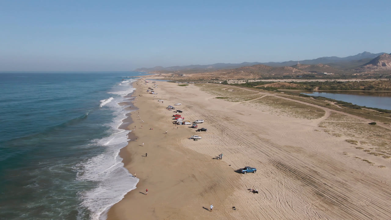 Aerial frame over Migrino, Baja California Sur showing ocean surf along the left edge, parked cars scattered on the sand, dunes, inland water, distant hills, and no visible people.