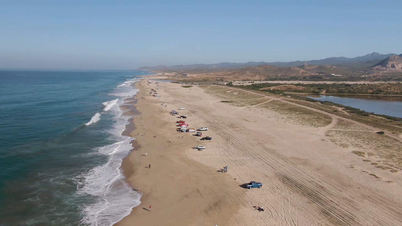 Aerial view in Migrino with a longer diagonal stretch of shoreline, white waves breaking beside tan sand, parked cars clustered near beach shade setups, inland water, hills, and no visible people.