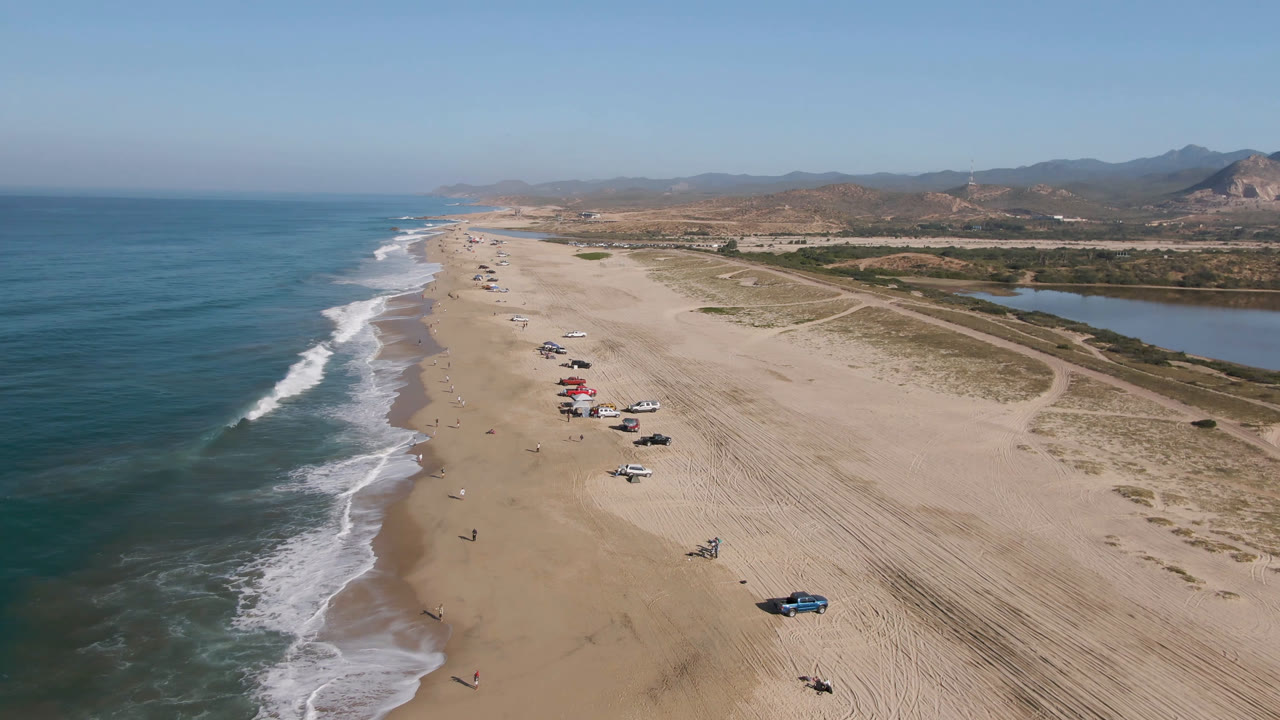 Wide aerial coastline with parked beach cars, surf, sand tracks, and hills.