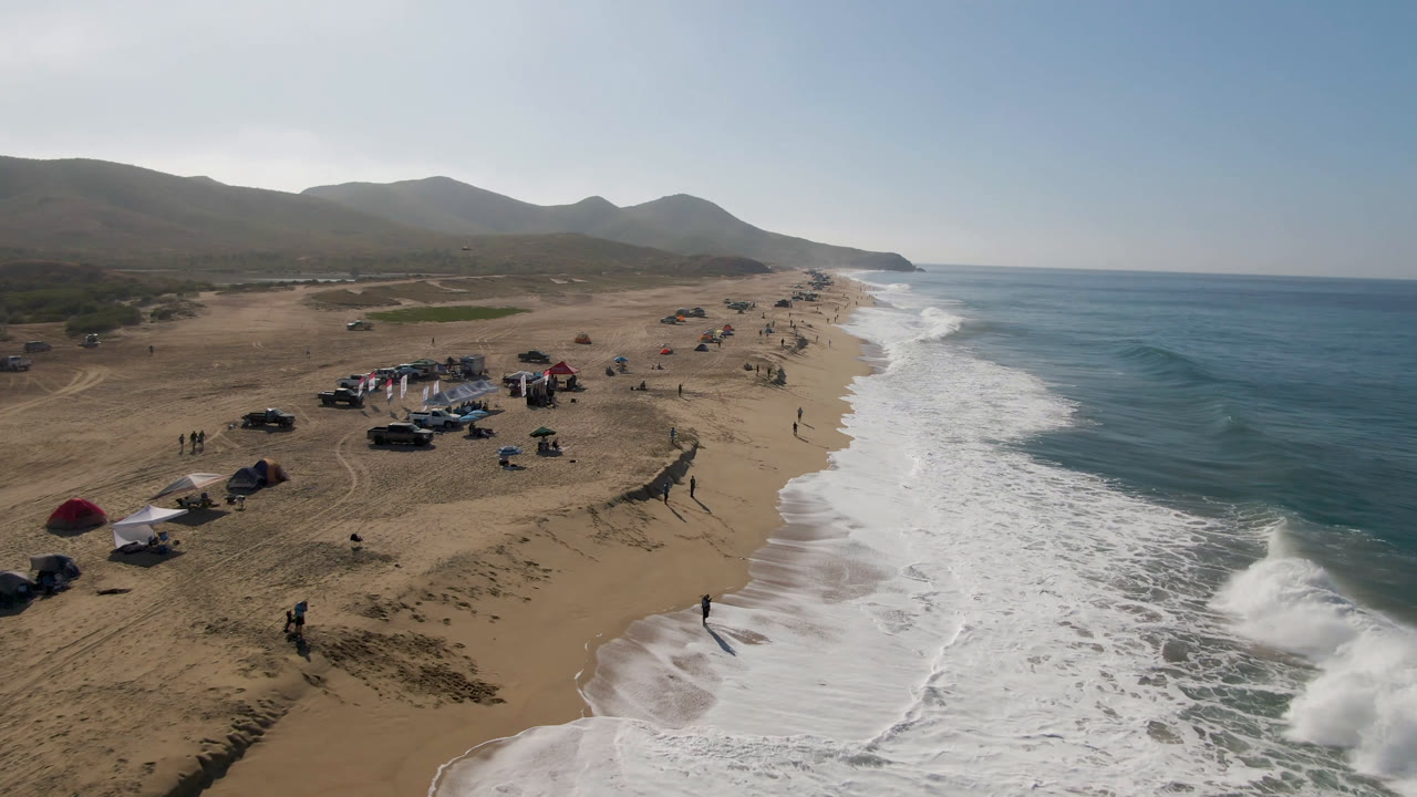 Aerial view of surf breaking beside a sandy beach with tents, vehicles, and people.
