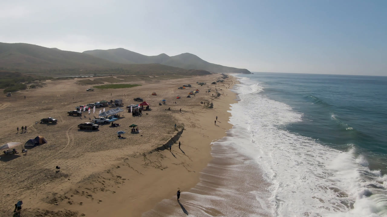 High aerial frame of Migrino beach showing surf washing up the right side, a group of tents and parked vehicles near the middle, and people spread across the sand under hazy daylight.