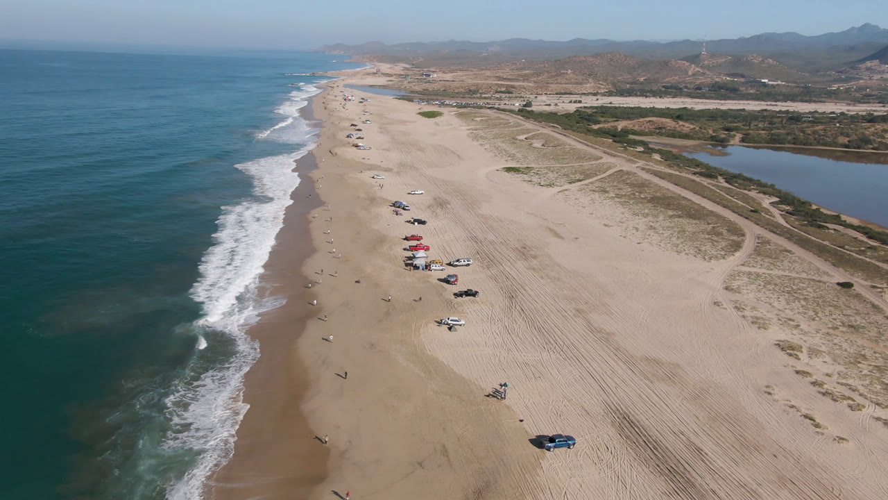 Aerial view of beach, surf, parked vehicles, lagoon, and dry hills.