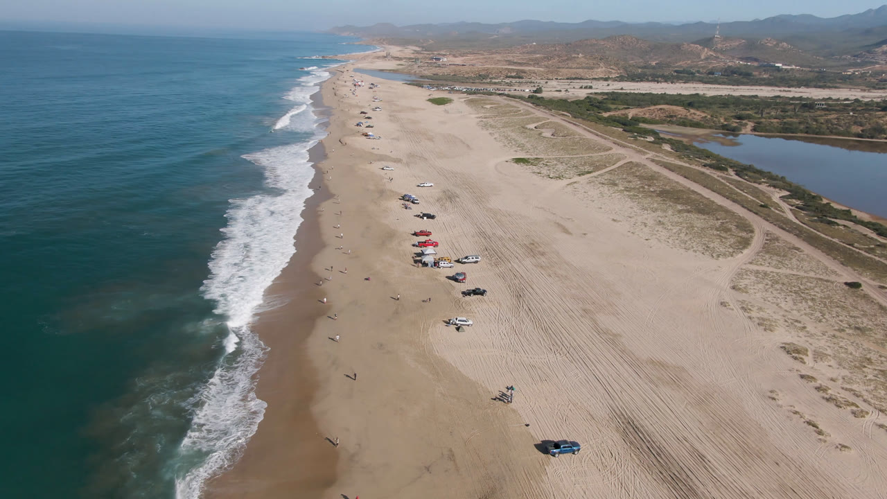 Aerial frame over Migrino showing the shoreline receding into the distance, white wave lines along the left side, vehicles grouped on the sand, lagoon water to the right, and no recognizable people visible.
