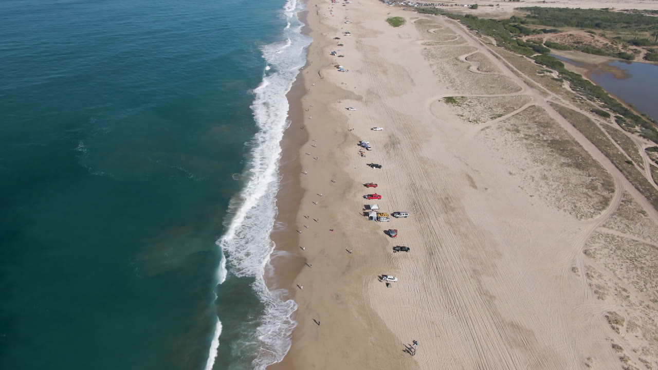 High aerial frame over Migrino, Baja California Sur, with teal ocean at left, white surf along the sand, parked cars and umbrellas near the beach center, and dune tracks inland.