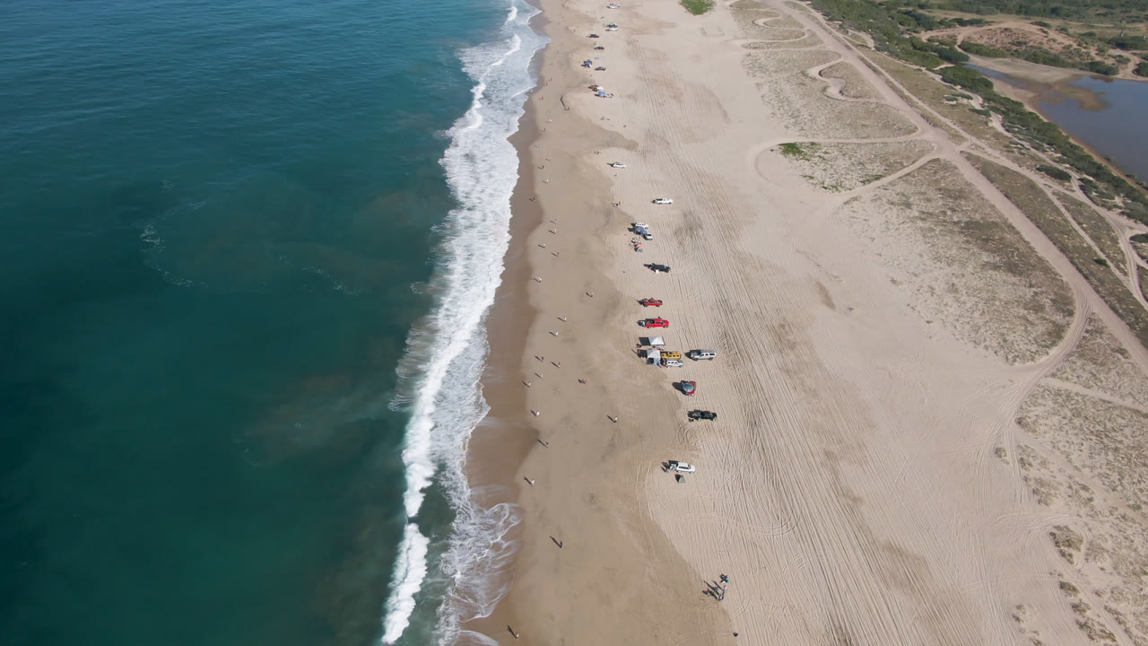 Aerial view in Migrino, Baja California Sur, showing a vertical line of breaking waves, broad tan beach, small beachgoers, clustered umbrellas, parked vehicles, and sandy paths through low dunes.