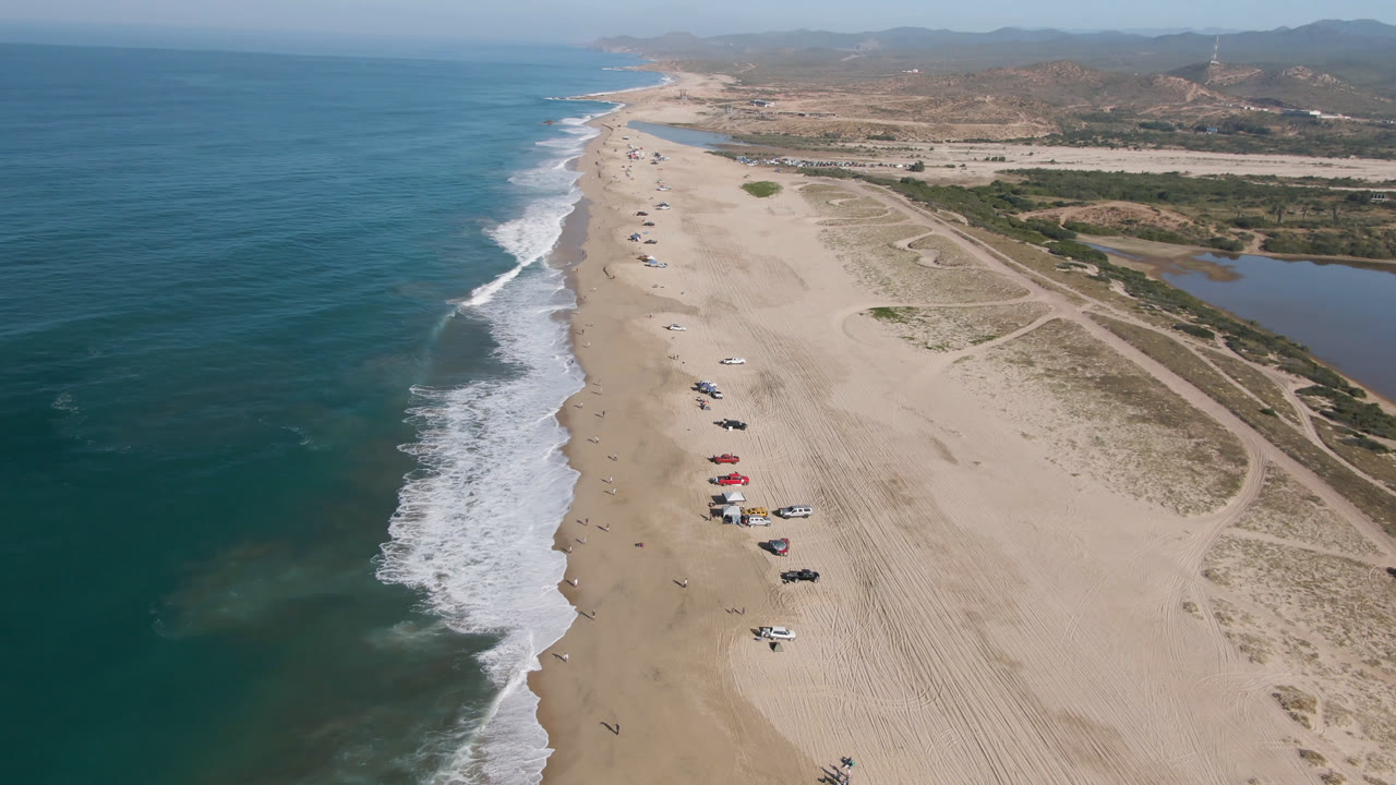 Aerial view of a sandy beach, ocean surf, parked vehicles, inland tracks, and low hills.