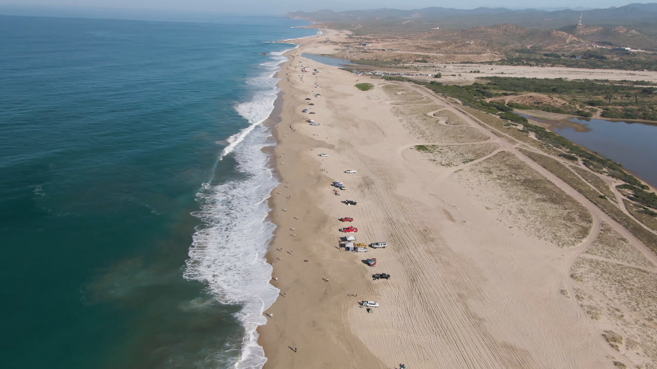 A second aerial frame over Migrino shows the surf line cutting diagonally through the beach, a loose row of vehicles farther down the sand, dirt paths inland, and no discernible people.