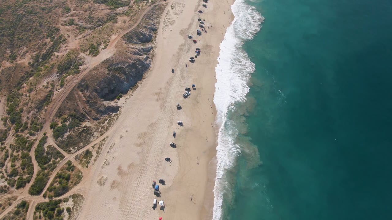 Overhead beach shoreline with dunes, surf, and small vehicles on sand.