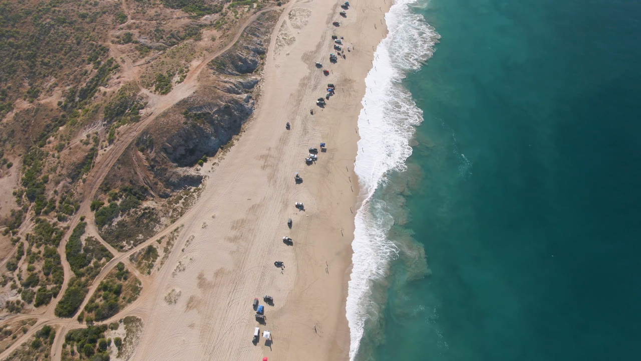 Drone view over Migrino beach with dune paths and brush on the left, scattered small vehicles on the sand, and a band of white surf separating beach from ocean.