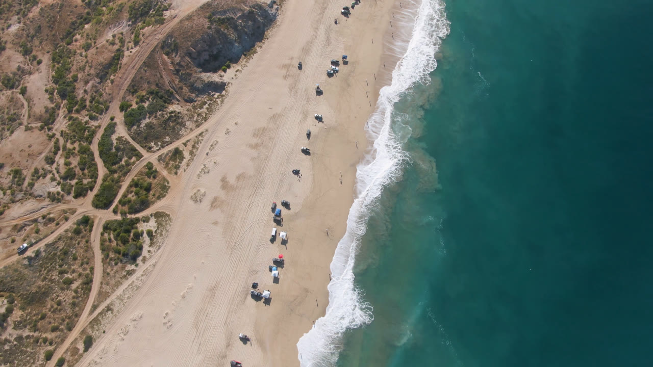 High aerial view of Migrino beach in Baja California Sur, Mexico, with dirt paths on the left, pale sand through the center, white surf on the right, and no discernible people visible.