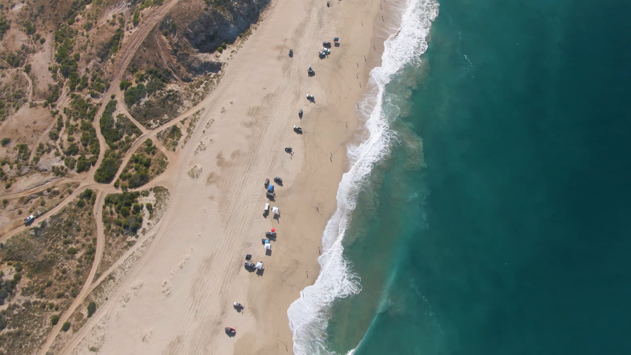 Overhead frame from Migrino, Baja California Sur, Mexico, showing umbrella clusters and parked vehicles along the sand, with the wave edge closer to the center and no discernible people visible.