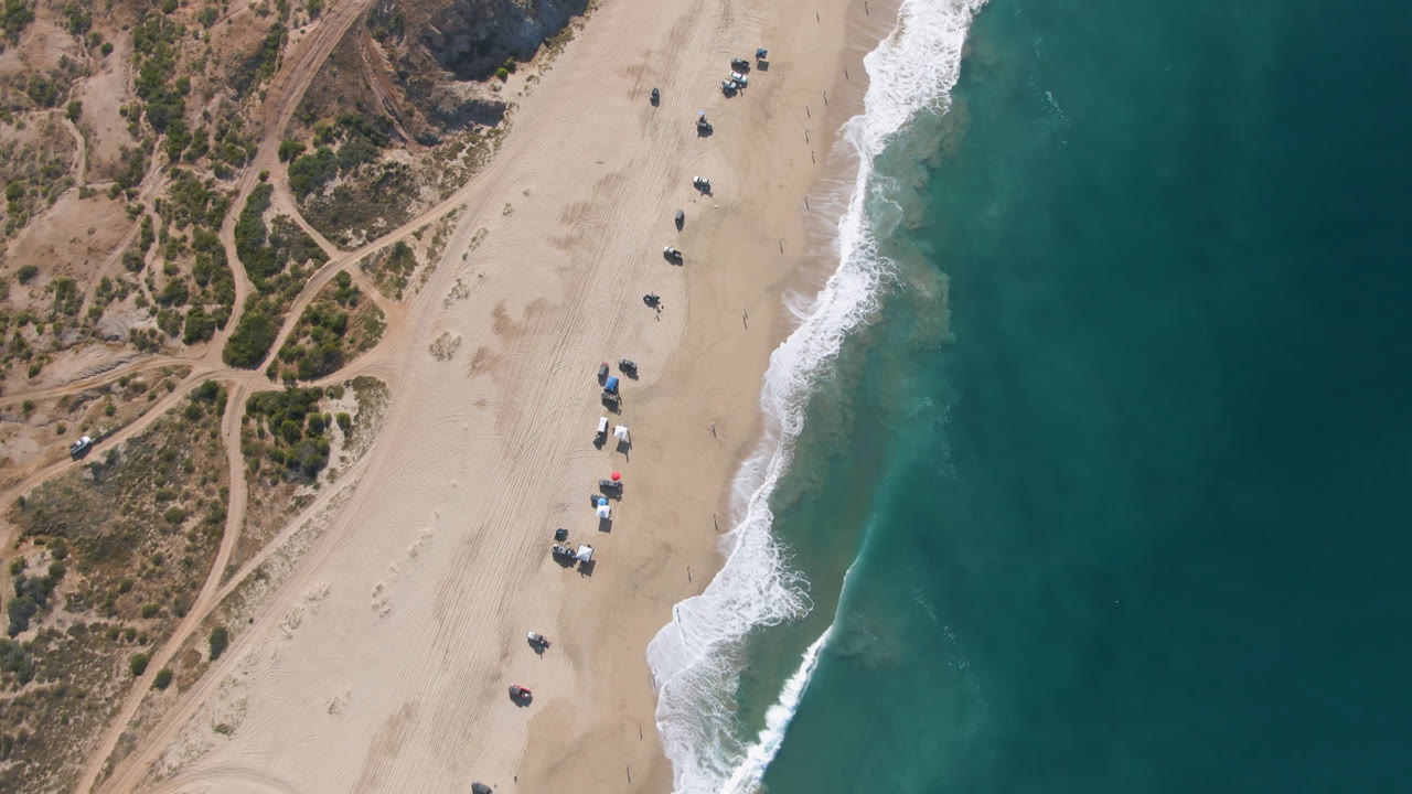 Top-down beach scene with waves, umbrellas, vehicles, sand, and dirt tracks.