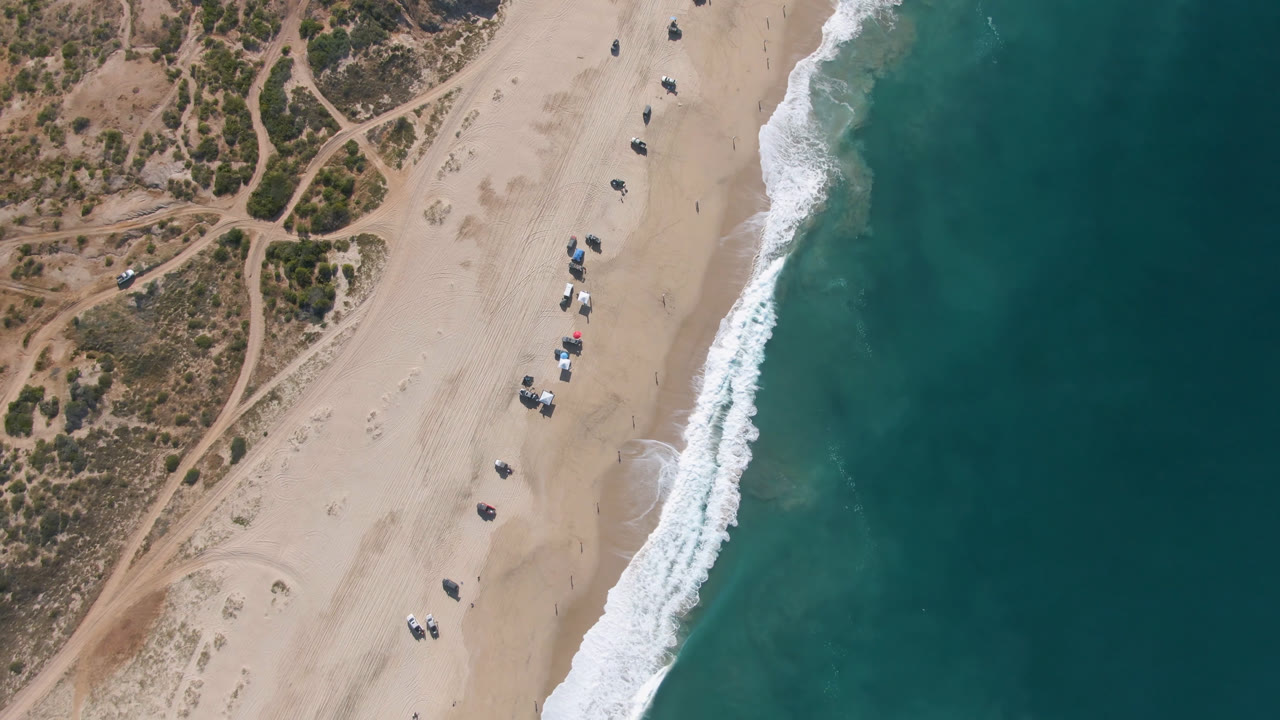 Overhead beach with surf, sand, dunes, and parked vehicles.