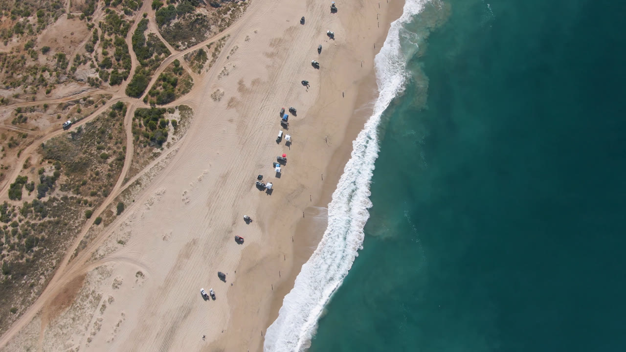 Aerial frame over Migrino beach showing the shoreline running diagonally, foam pushing onto sand, dune paths at upper left, parked vehicles, and small distant beachgoers.