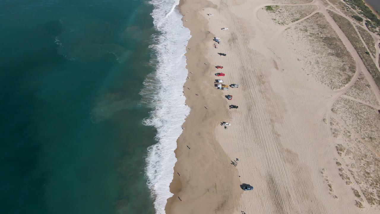 Overhead beach with ocean surf, umbrellas, vehicles, and sand tracks
