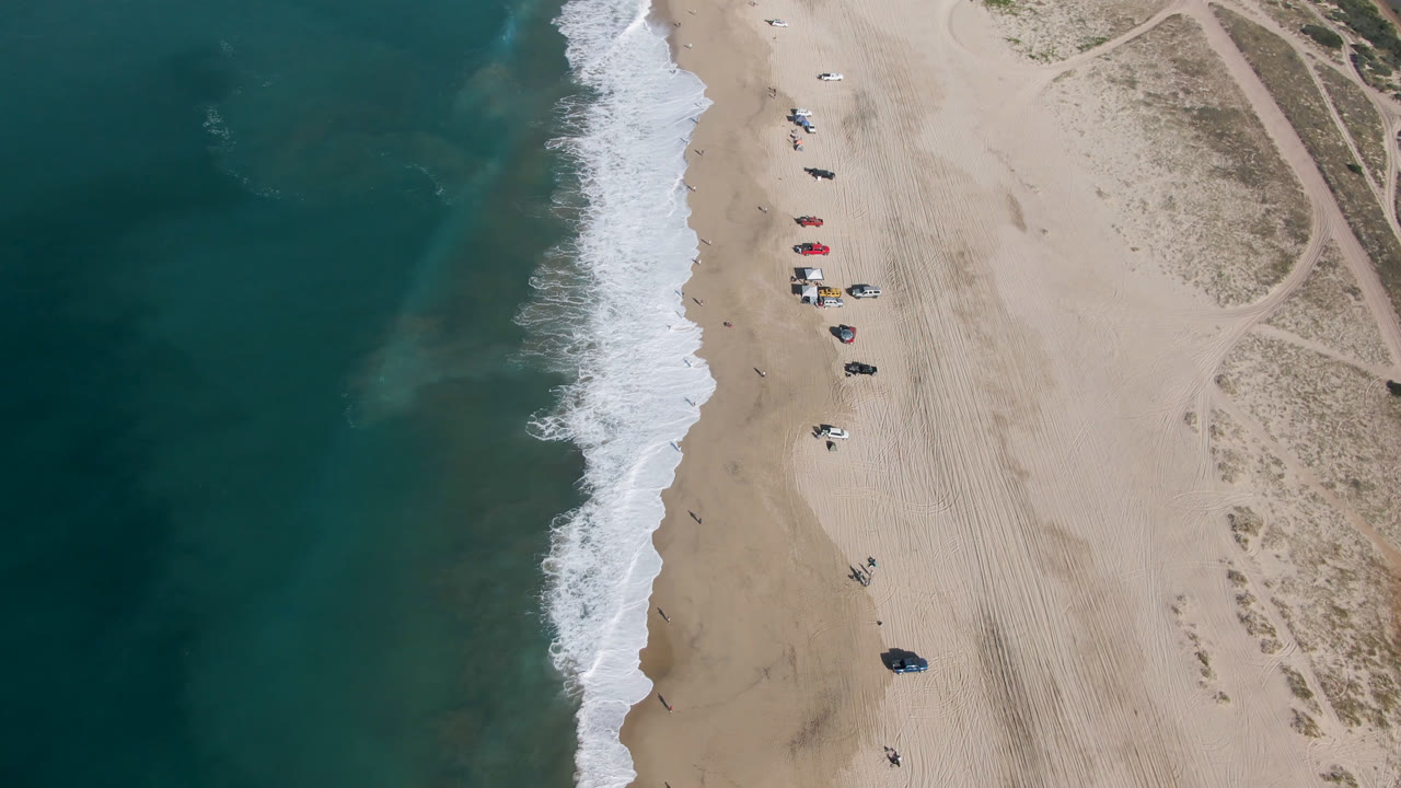Aerial frame over Migrino beach showing the surf edge slightly farther along the sand, with clustered umbrellas and vehicles near the waterline and looping dune paths to the right.