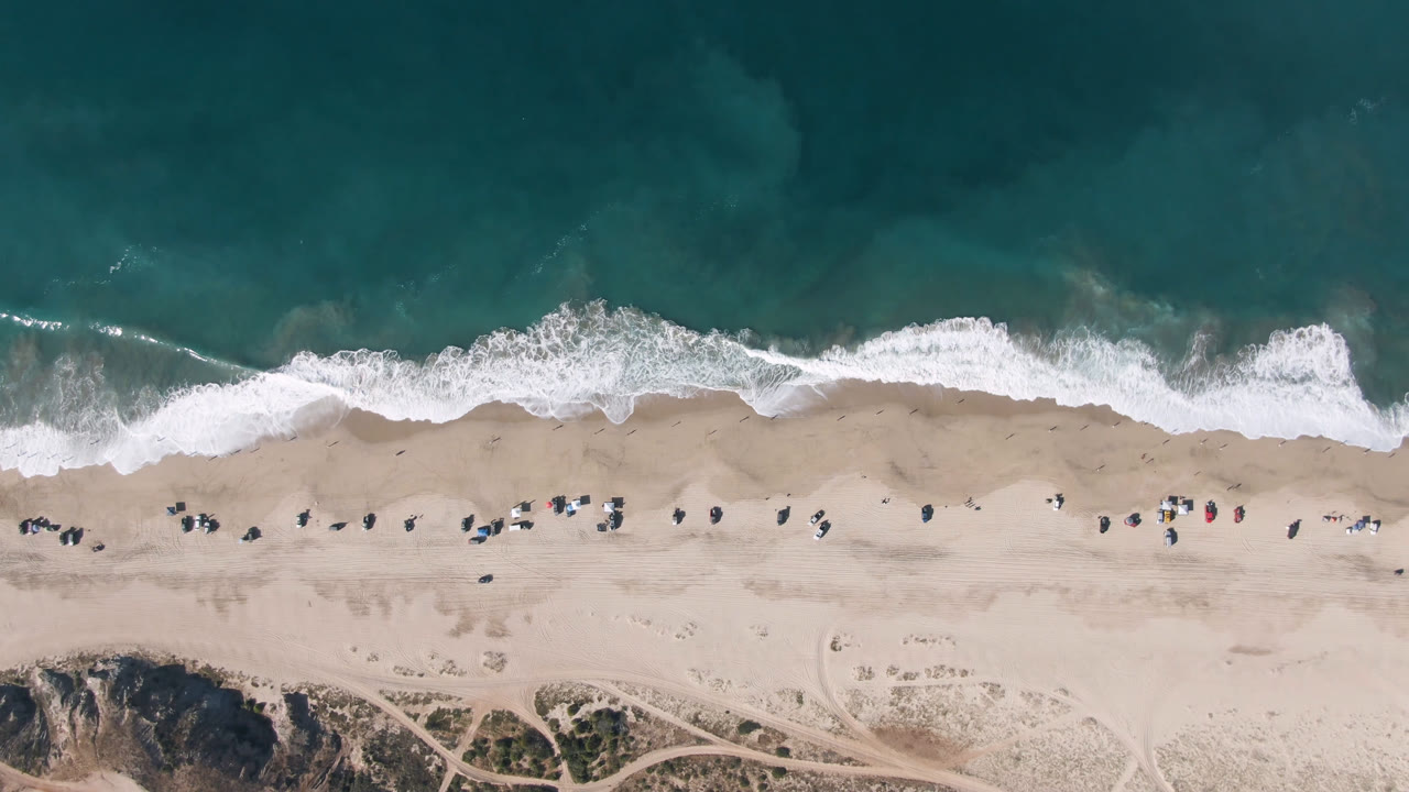 Overhead view of Migrino beach in Baja California Sur with teal ocean at the top, white surf crossing the sand, tiny beachgoers, and dune tracks along the lower edge.