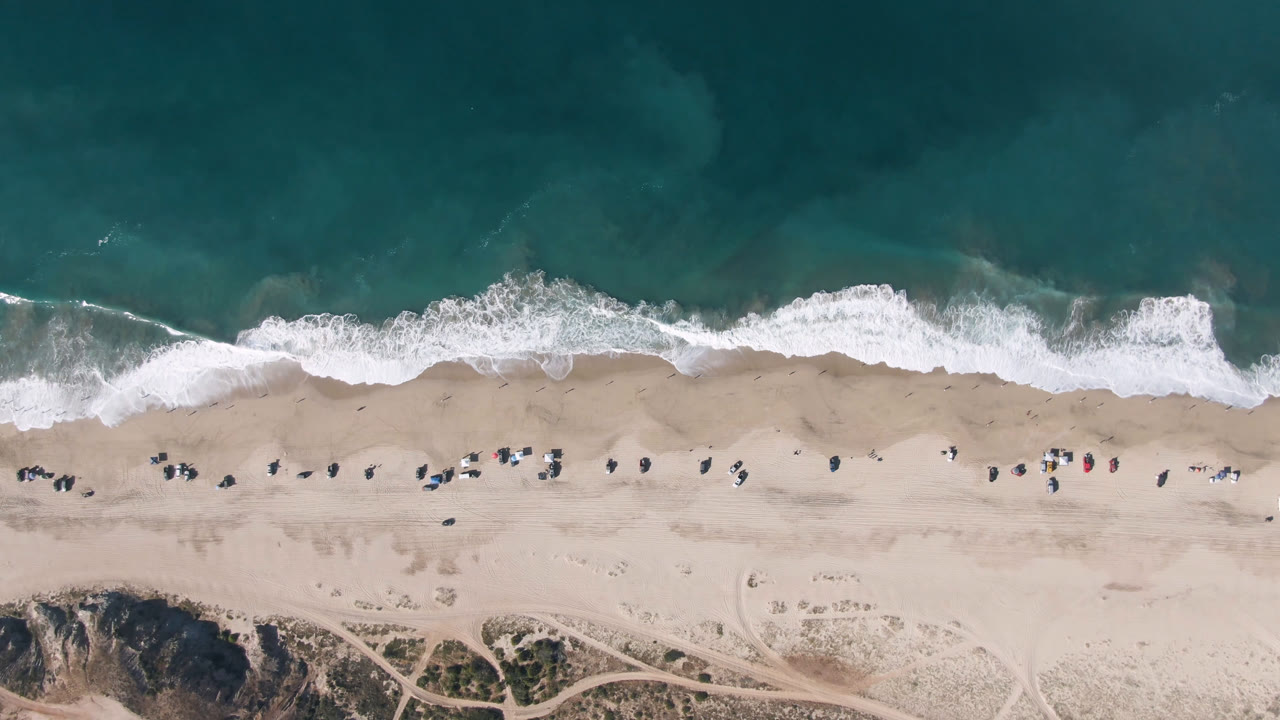 Aerial frame from Migrino showing a long strip of pale sand, uneven white wave foam, small people and beach items near the waterline, and scrubby dunes below.