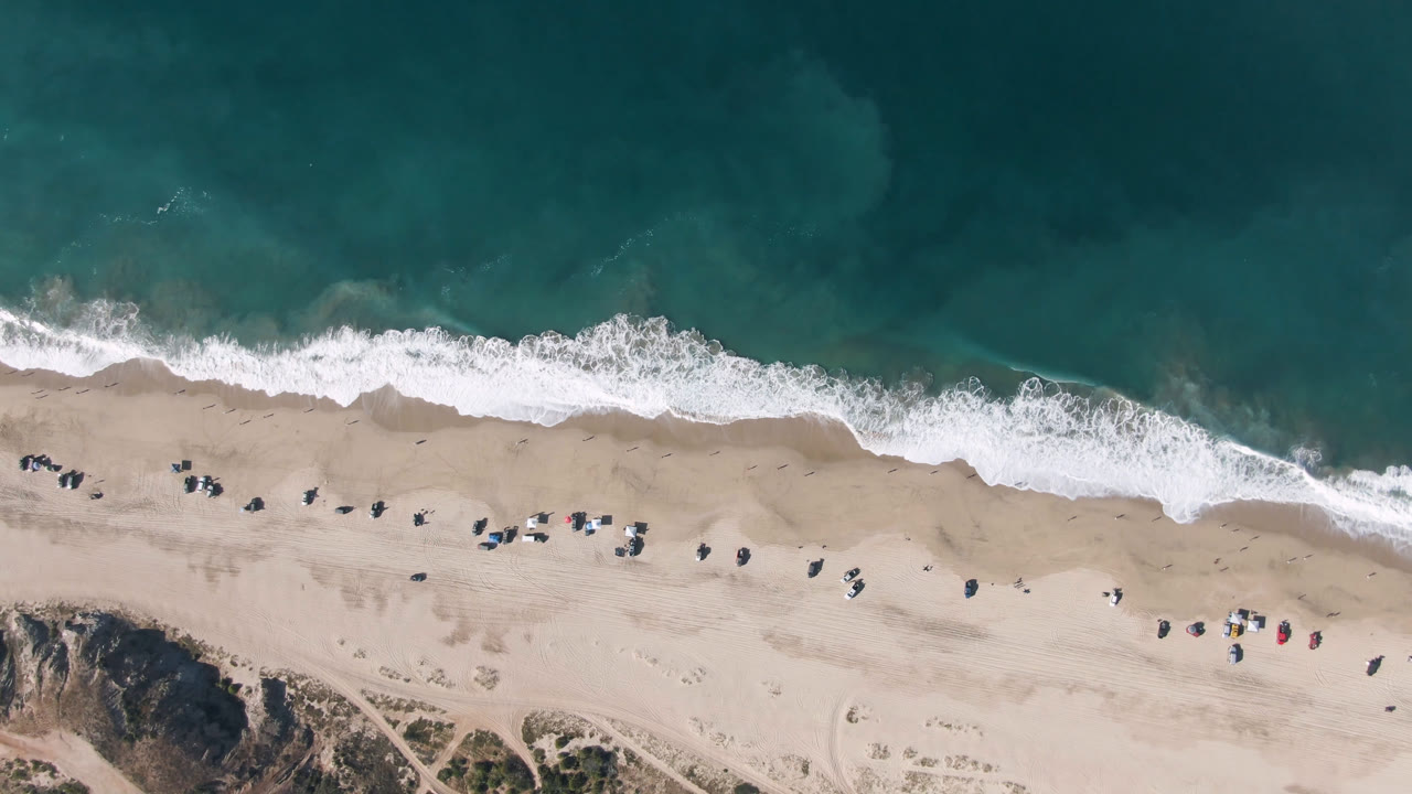 Overhead shoreline with teal water, white surf, pale sand, and scattered tiny beach objects.