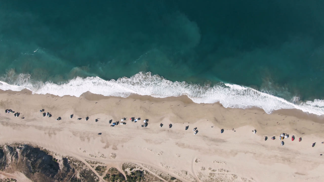 Overhead aerial frame in Migrino showing a slightly shifted surf band across the beach, open sand below it, bluff textures at the bottom, and no recognizable people visible.
