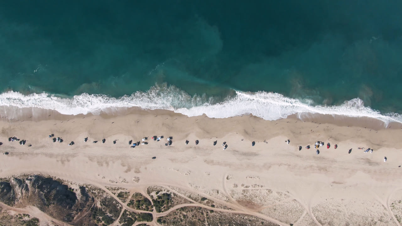 Overhead beach shoreline with teal water, breaking surf, pale sand, and dune paths.