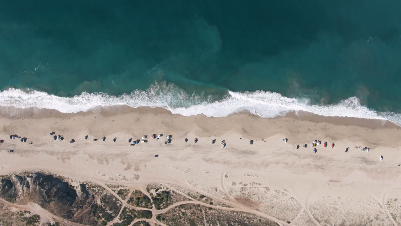 Aerial frame over Migrino beach showing a fresh line of white surf, broad dry sand, small distant beach figures or objects, and dune tracks along the lower edge.