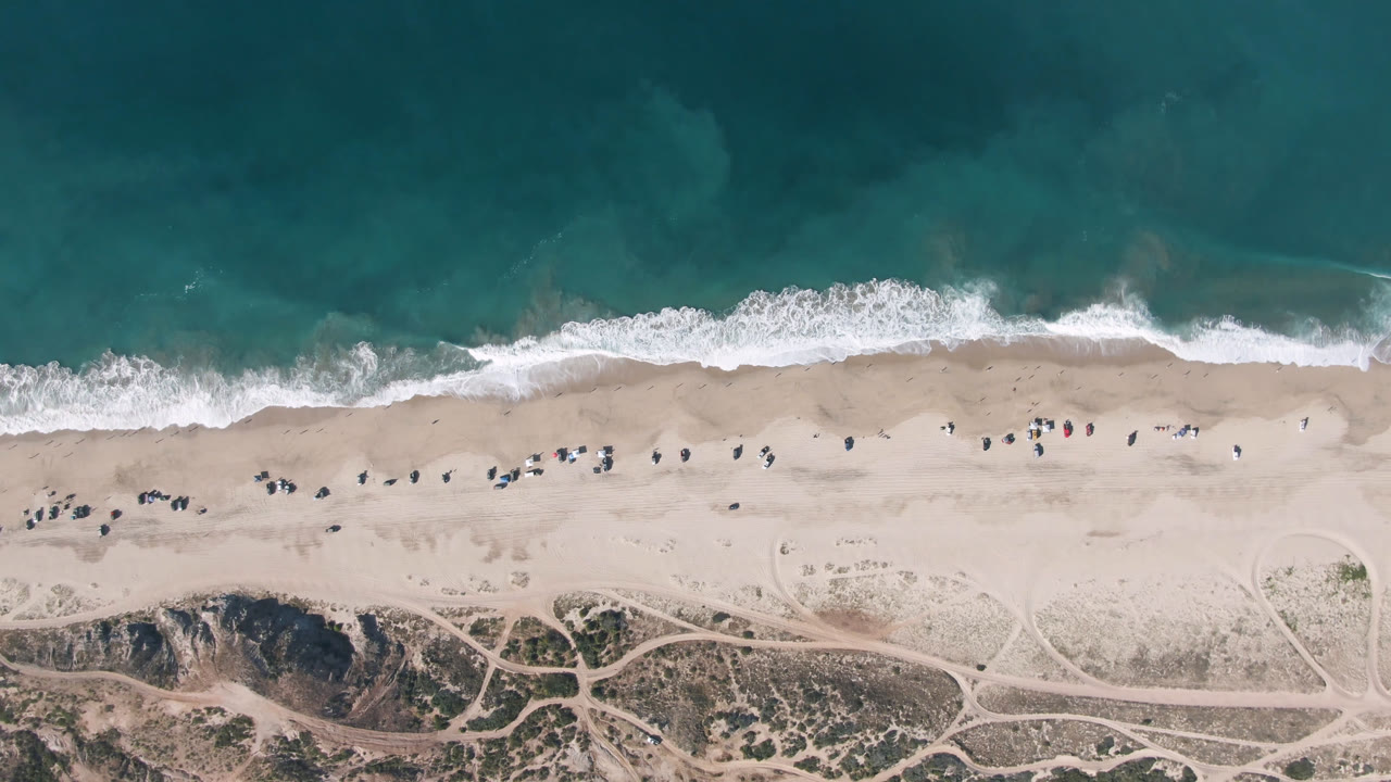 Overhead frame from Migrino, Baja California Sur showing teal water, a white surf line, pale sand, dune tracks, and tiny visible beachgoers near the shoreline.