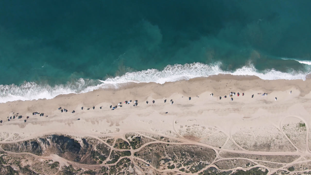 Aerial view of the Migrino shoreline with waves slightly farther across the sand, scattered tiny people or beach objects, and intersecting tracks in the dune area below.