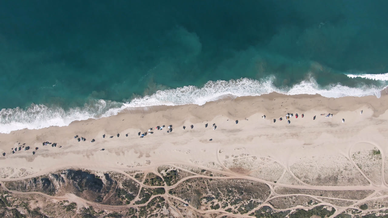 Top-down coast with teal water, foamy waves, sandy beach, and sparse beach activity.