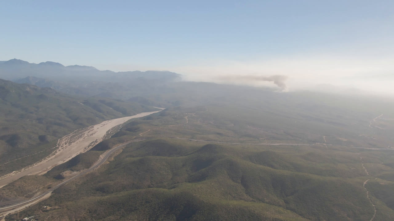 Aerial frame over Migrino, Baja California Sur showing green-brown hills, a pale riverbed crossing the valley, thin roads, distant mountains, horizon smoke, and no visible people.