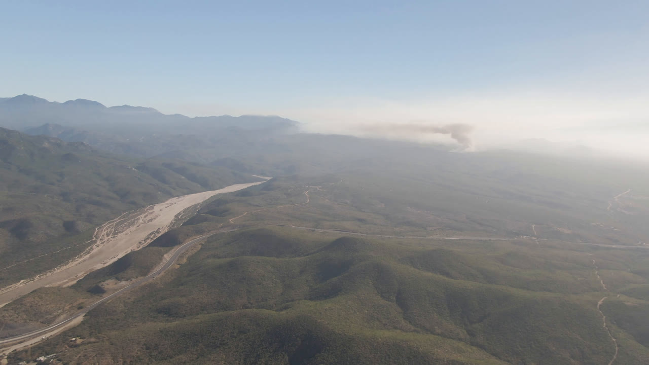A high aerial view in Migrino, Baja California Sur with rounded foreground hills, a light-colored riverbed bending through the valley, road cuts, haze, distant smoke, and no visible people.