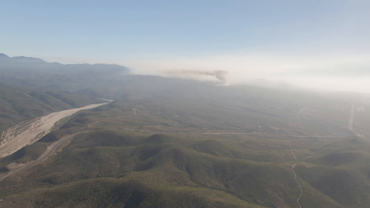 Aerial mountain valley with a pale riverbed and distant smoke plume.