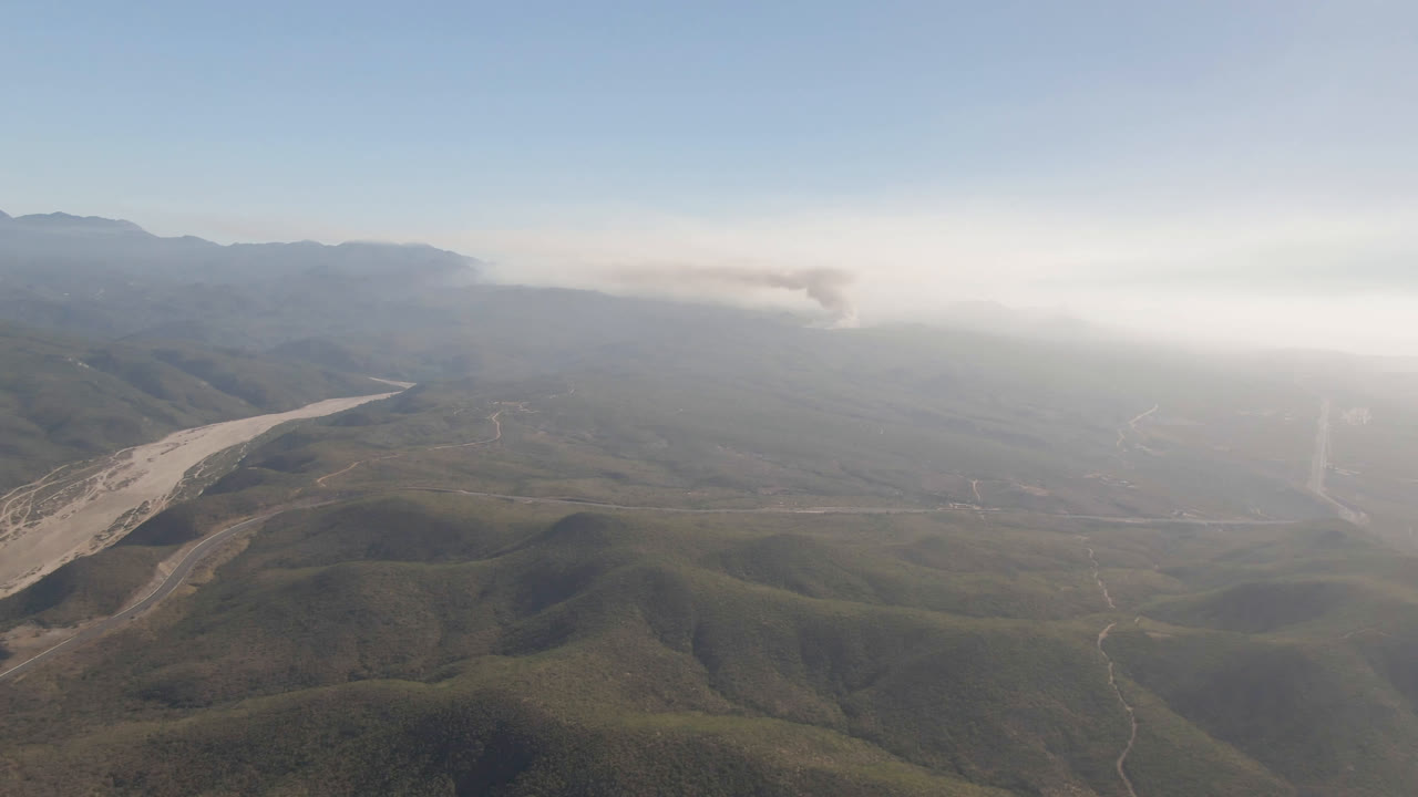 Wide aerial frame over Migrino mountain ridges with the pale valley drainage visible left of center, a hazy background, distant smoke, and no visible people.