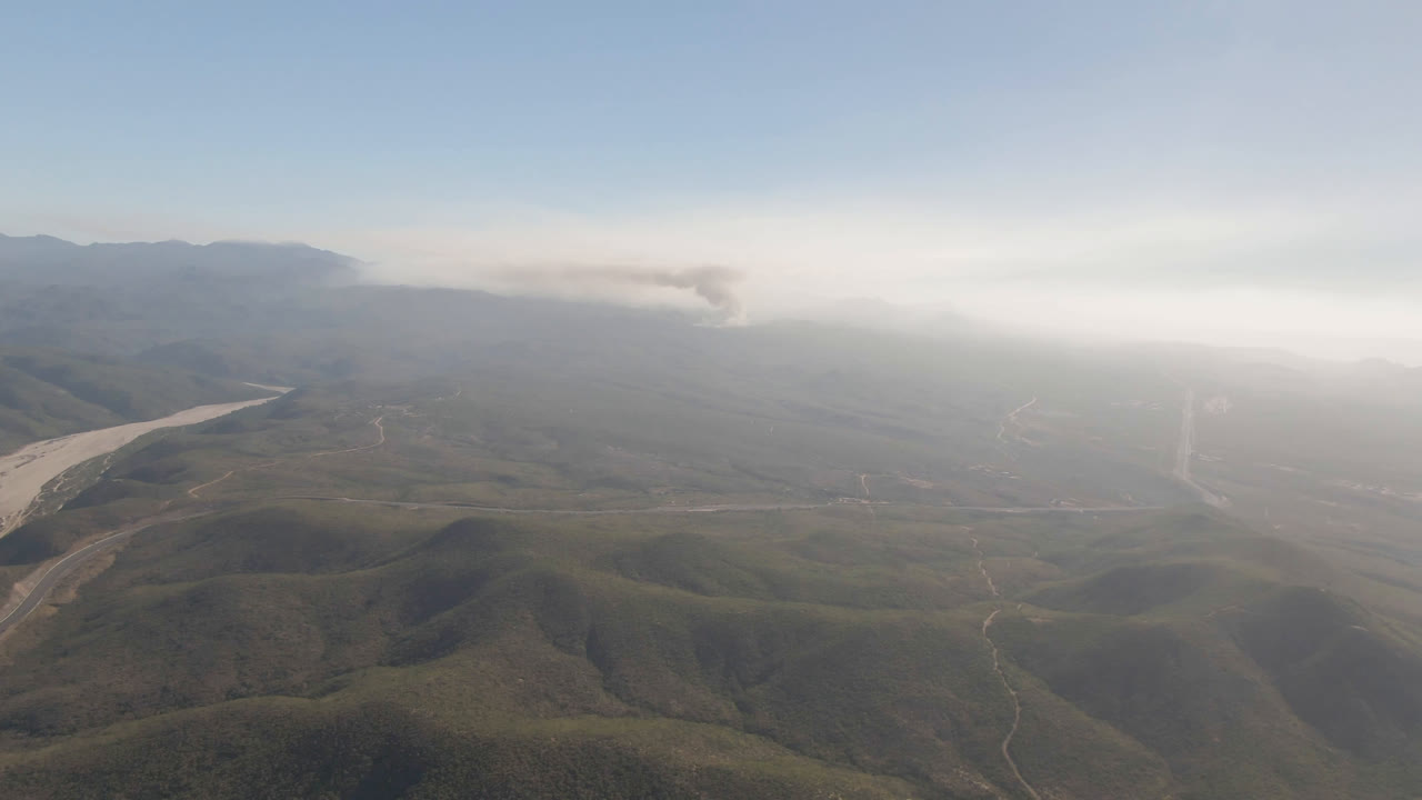 Opening frame over Migrino, Baja California Sur, with green-brown ridges in the foreground, a pale river channel at left, haze in the valley, distant smoke, and no visible people.