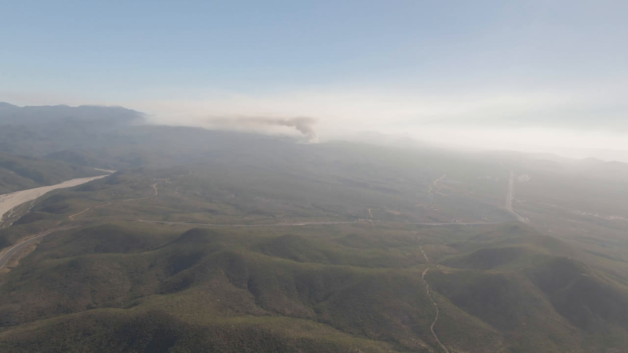 Second frame in Migrino, Baja California Sur, looking across rounded hills and faint dirt roads toward a smoke plume spreading along the distant horizon, with no visible people.