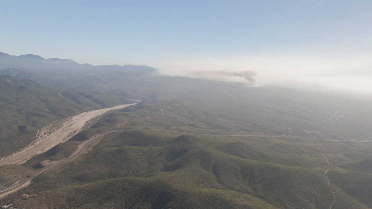 Wide aerial view over Migrino, Baja California Sur, with green-brown ridges, a pale river wash bending through the valley, distant haze on the horizon, and no visible people.