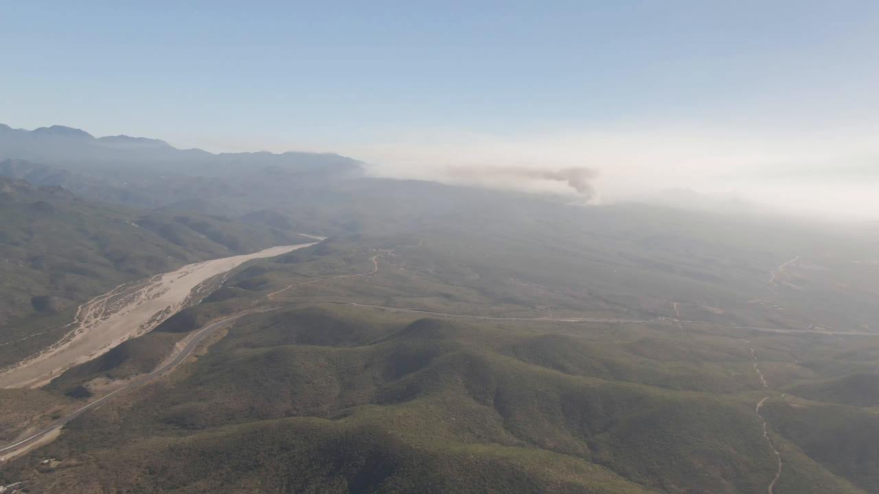 Aerial frame over Migrino, Baja California Sur, showing the pale wash more centrally between rounded hills, with layered mountains and a bright smoke or cloud bank in the background, and no visible people.